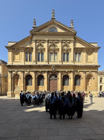 Oxford graduands lined up waiting to go into the Sheldonian Theatre on a sunny day with a clear blue sky. The graduands are wearing gowns and hoods