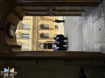 Img Looking through a doorway at Oxford graduands lined up waiting to go into the Sheldonian Theatre. The graduands are wearing gowns and hoods